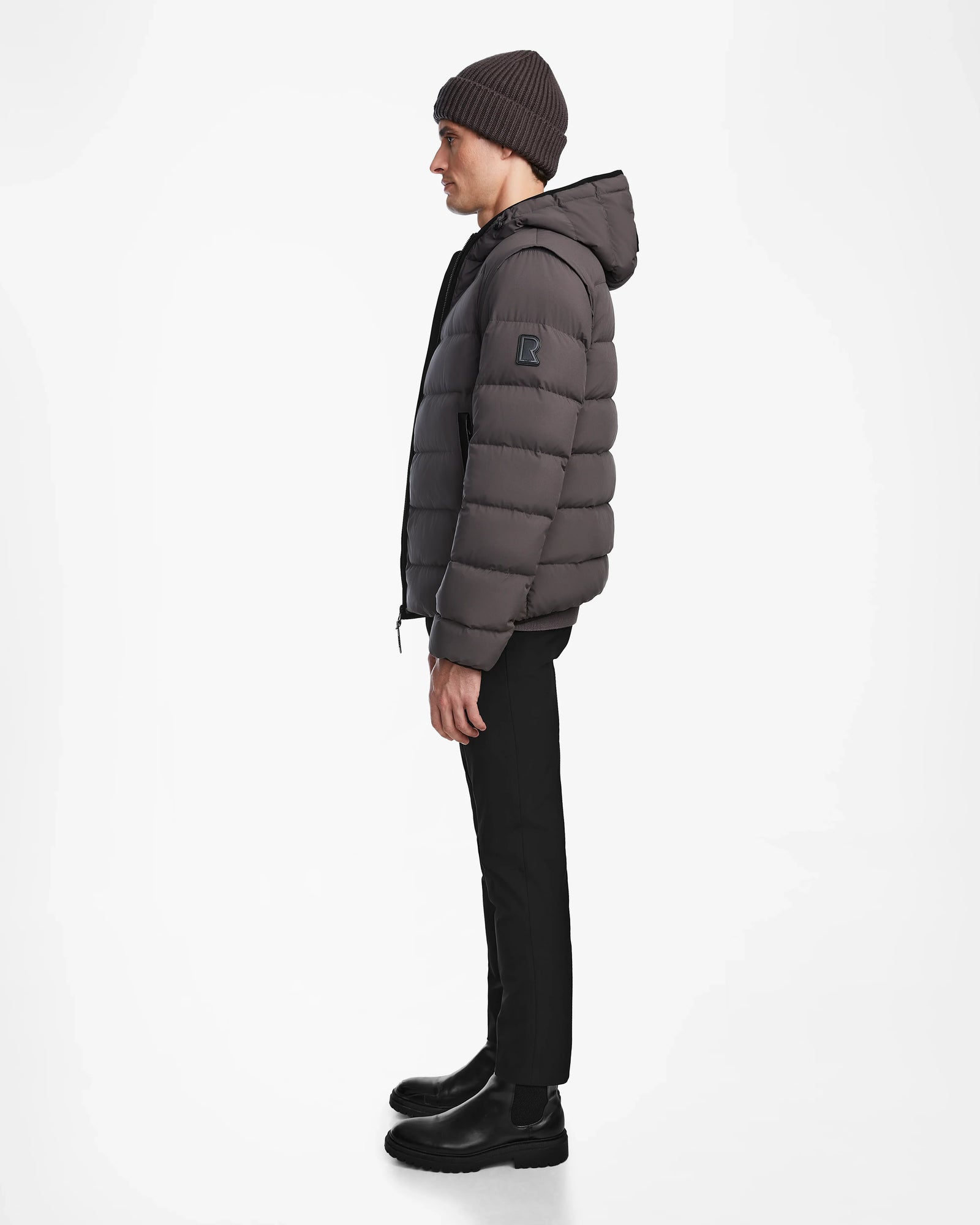 A man stands in profile against a white background, wearing the Rudsak DEMIAN M-R - M123504 Grey mens down puffer jacket with a hood, paired with black pants, black boots, and a dark knit beanie.