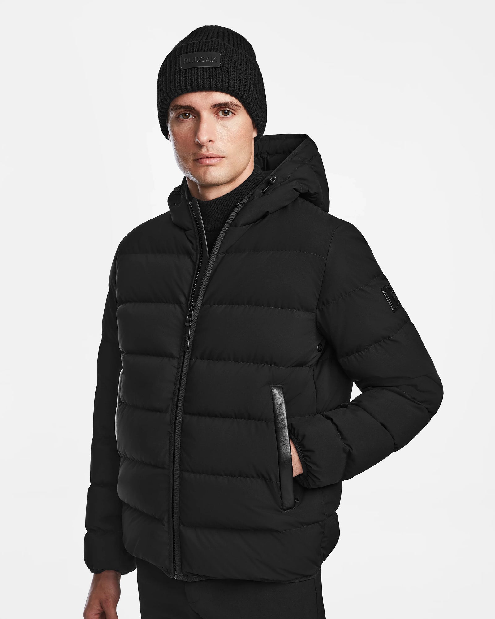 A man stands facing forward with one hand in his pocket, wearing the Rudsak DEMIAN M - M123507 Black men’s down puffer jacket with a hood and a black knit beanie, against a plain white background.