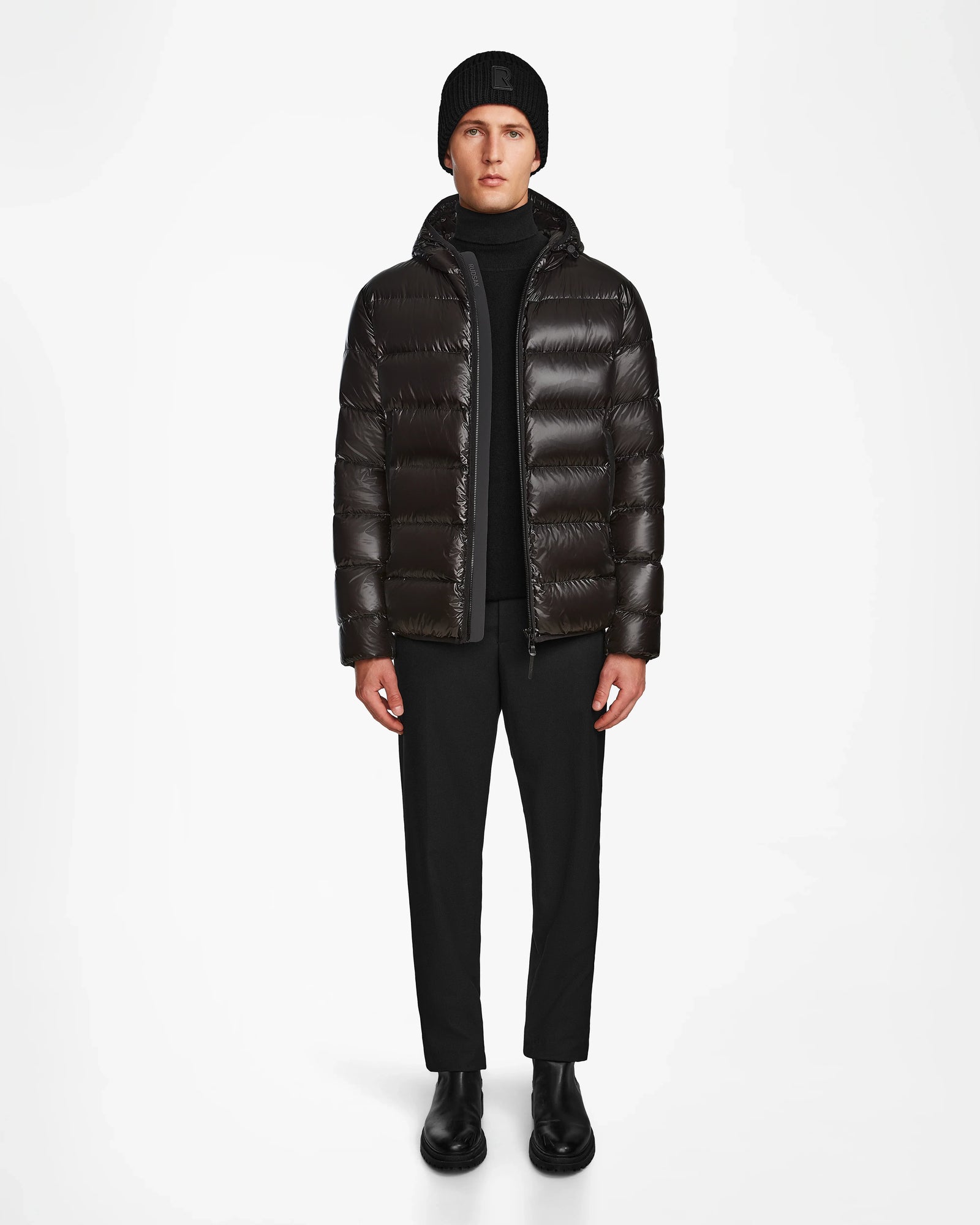 A man faces forward wearing the Rudsak DEMIAN S - M123508 Black mens down puffer, paired with a black beanie, black pants, and boots against a plain white background.
