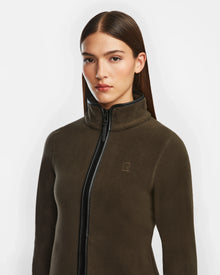 A woman with straight brown hair wears the Rudsak EDITH - 8524617 Army zip-up sweater in dark olive, featuring a high collar, subtle chest logo, soft polar fleece, and vegan leather trim, standing against a plain white background.