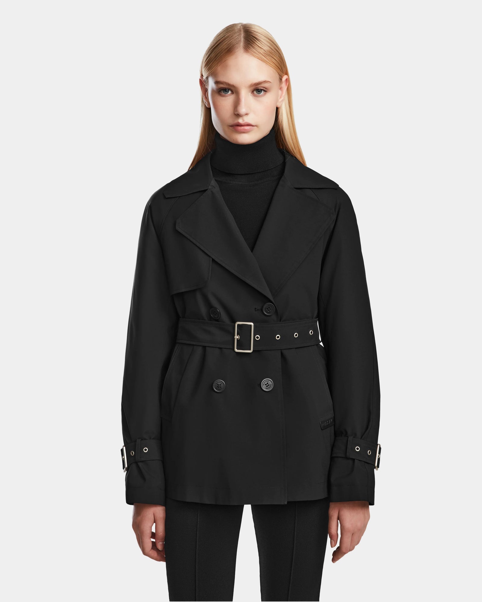 A woman with long blonde hair models the Rudsak ELENOR - 8124707 Black womens trench coat with a shearling collar and black belt over a black turtleneck and pants, standing against a plain white background.
