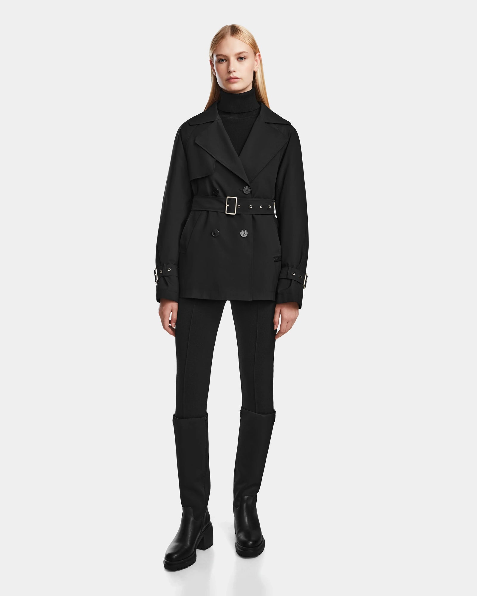 A woman faces forward against a plain light background, wearing the Rudsak ELENOR - 8124707 Black trench coat with a shearling collar, paired with a black turtleneck, fitted pants, and knee-high boots.