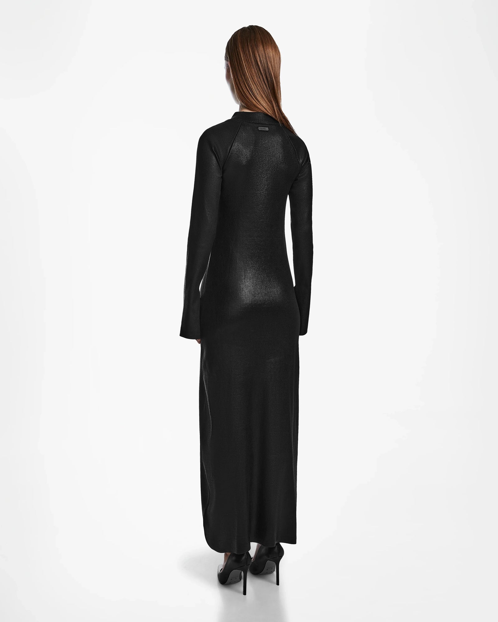 A woman with long brown hair faces away, wearing the Rudsak FAIZA - 8524579 Black mock-neck maxi dress and black heels, showcasing contemporary style against a plain white background.