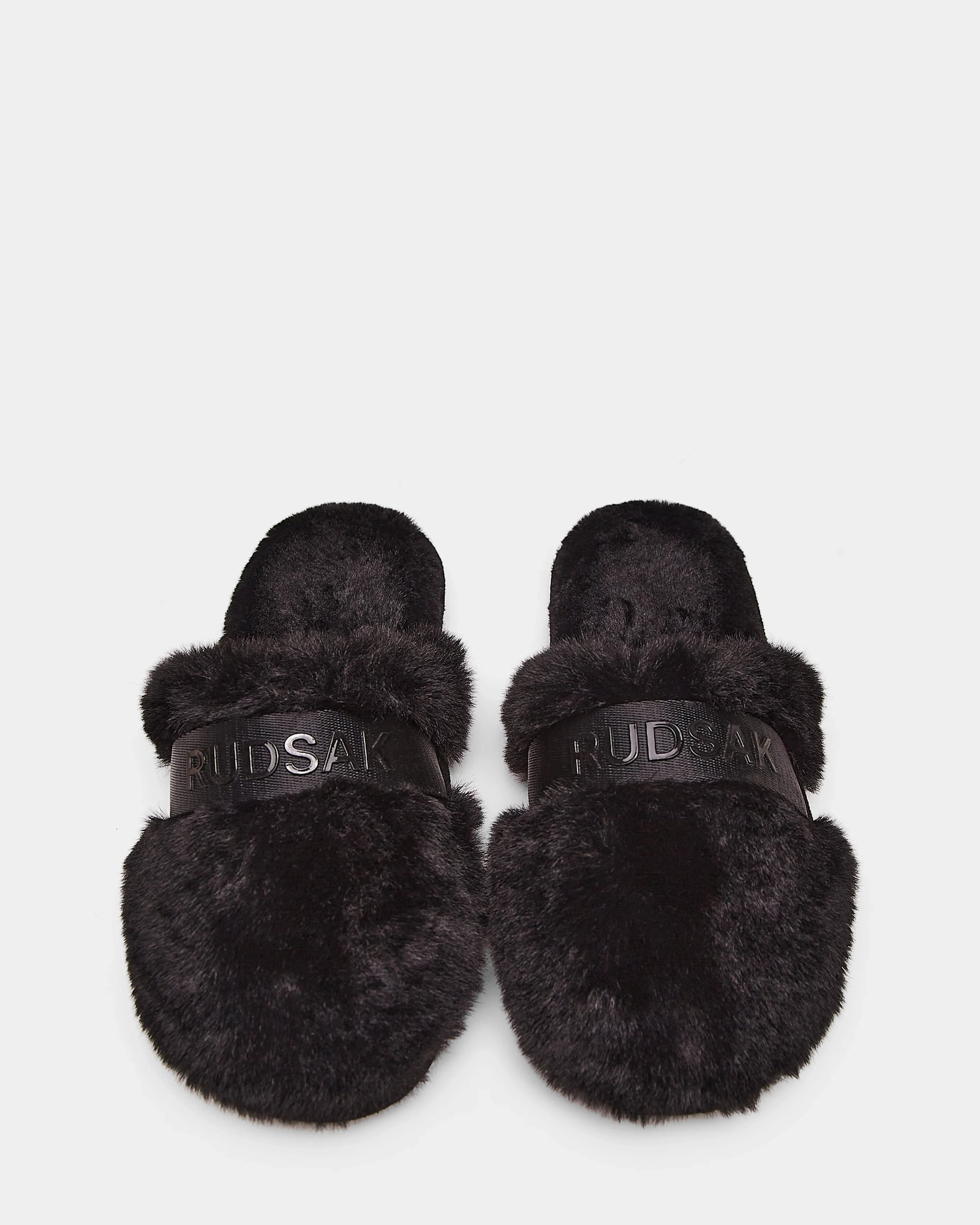 The Rudsak FLUFF - L223509 Black slippers feature cozy black faux-fur and RUDSAK branding across the straps, shown on a white background.