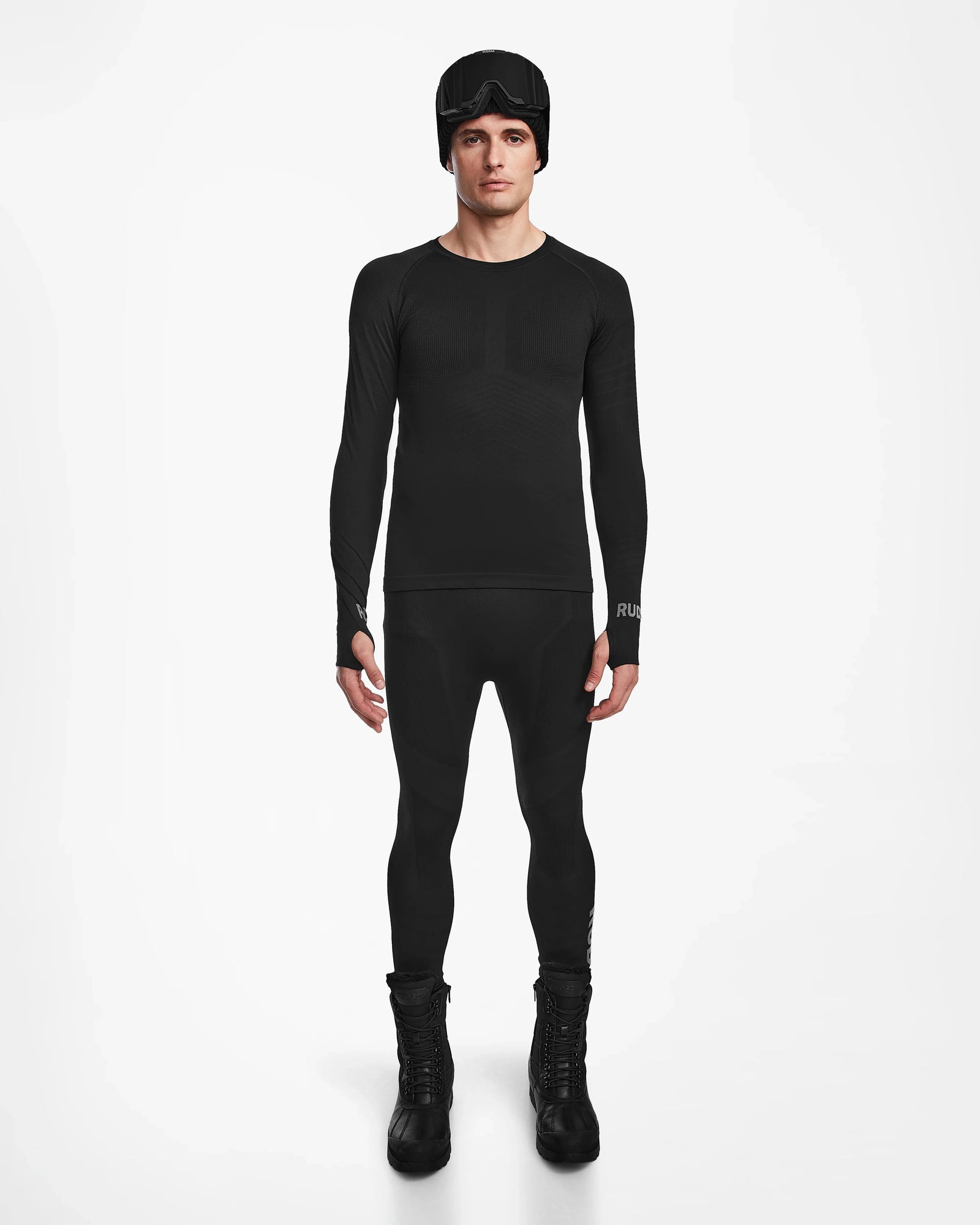 A man stands facing forward wearing the Rudsak FRANC - 6524541 Black fitted men’s thermal set with matching leggings, tall boots, and a black beanie with goggles, posed against a plain white background.