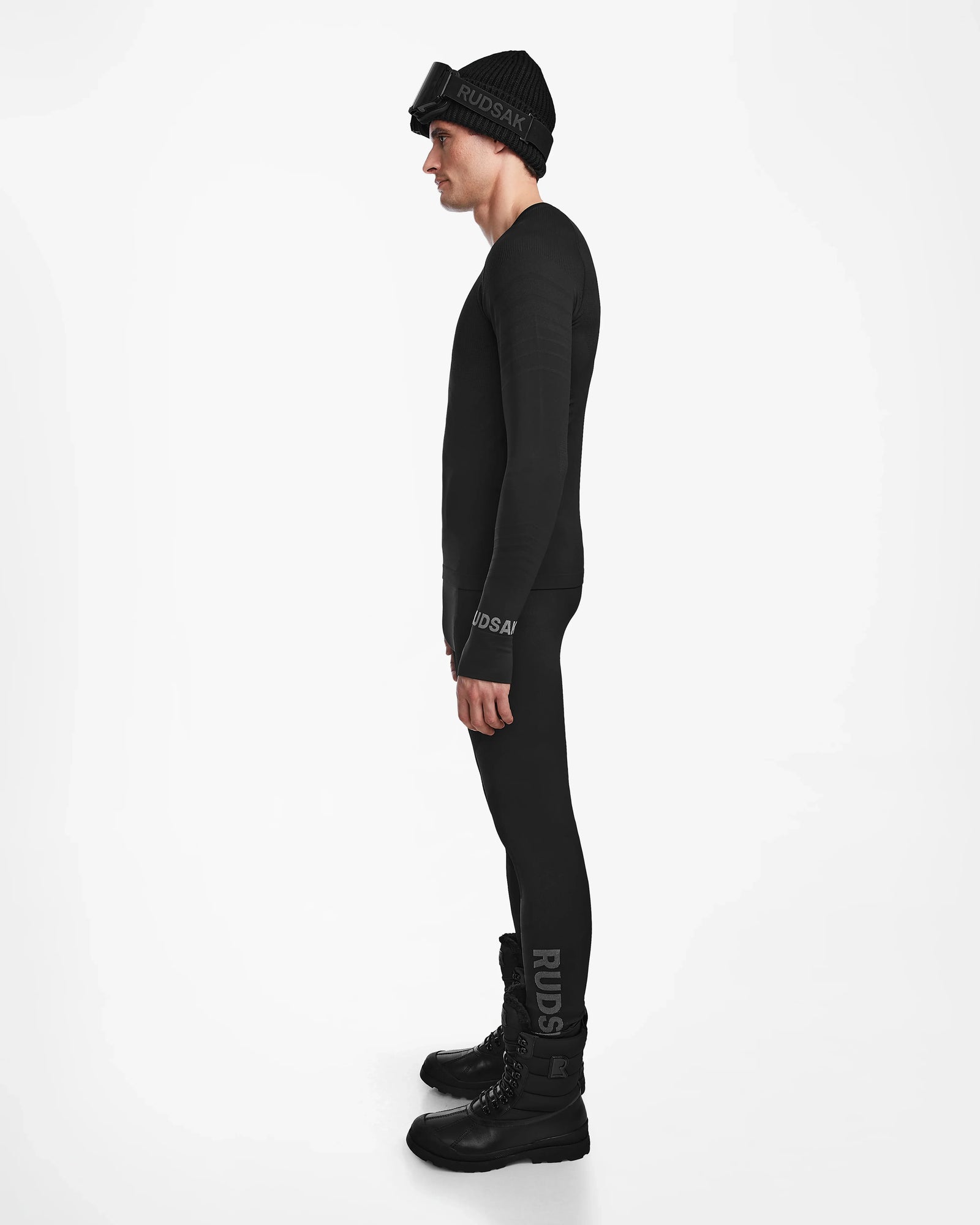 A man stands in profile against a white background wearing the Rudsak FRANC - 6524541 Black men’s thermal set, paired with a black Rudsak beanie, ski goggles, and boots. Both the clothing and beanie feature the Rudsak logo.