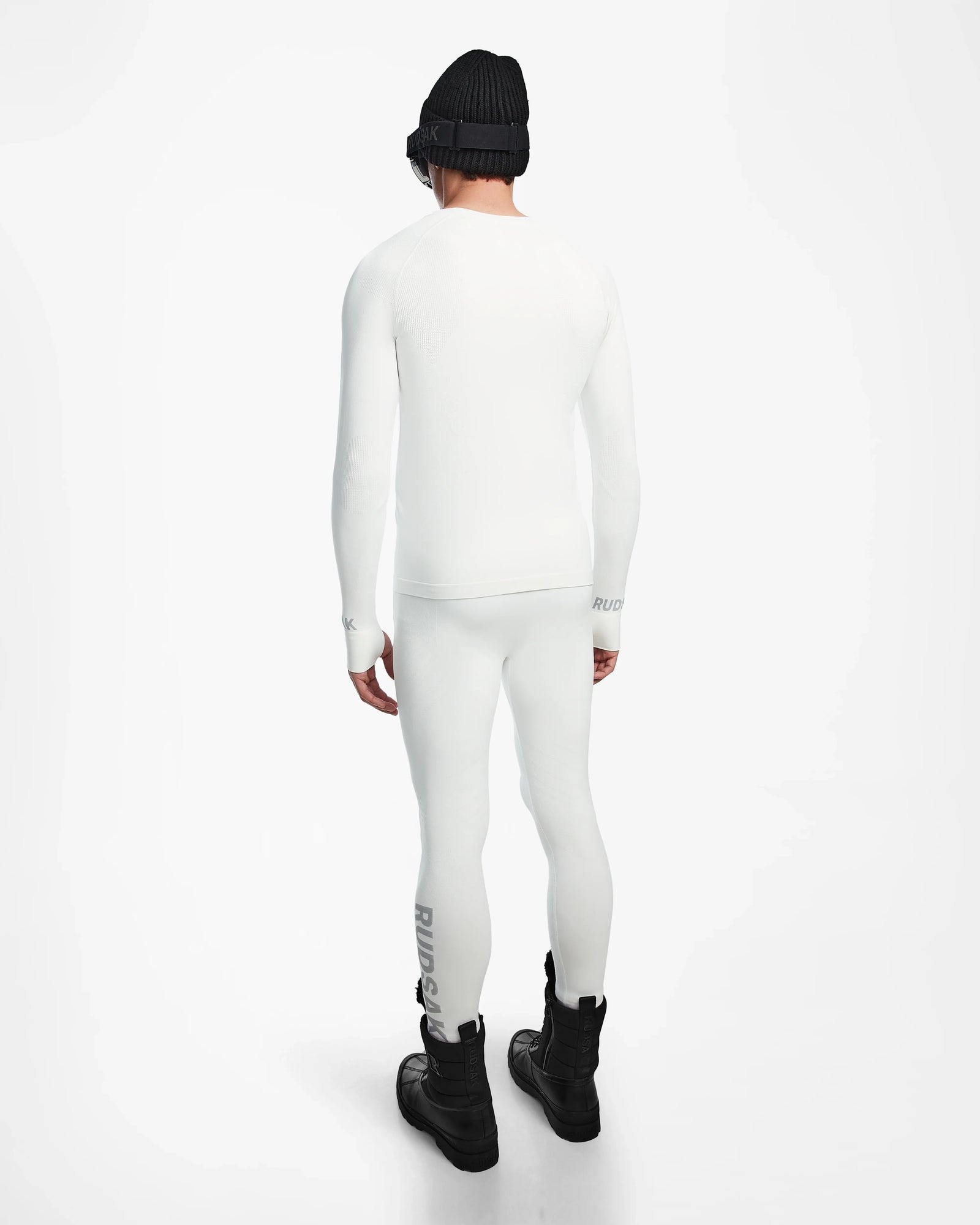 A person faces away, wearing the Rudsak FRANC - 6524541 Iceland white stretch base layer top and leggings, black winter boots, a black beanie, and black goggles against a plain white background.