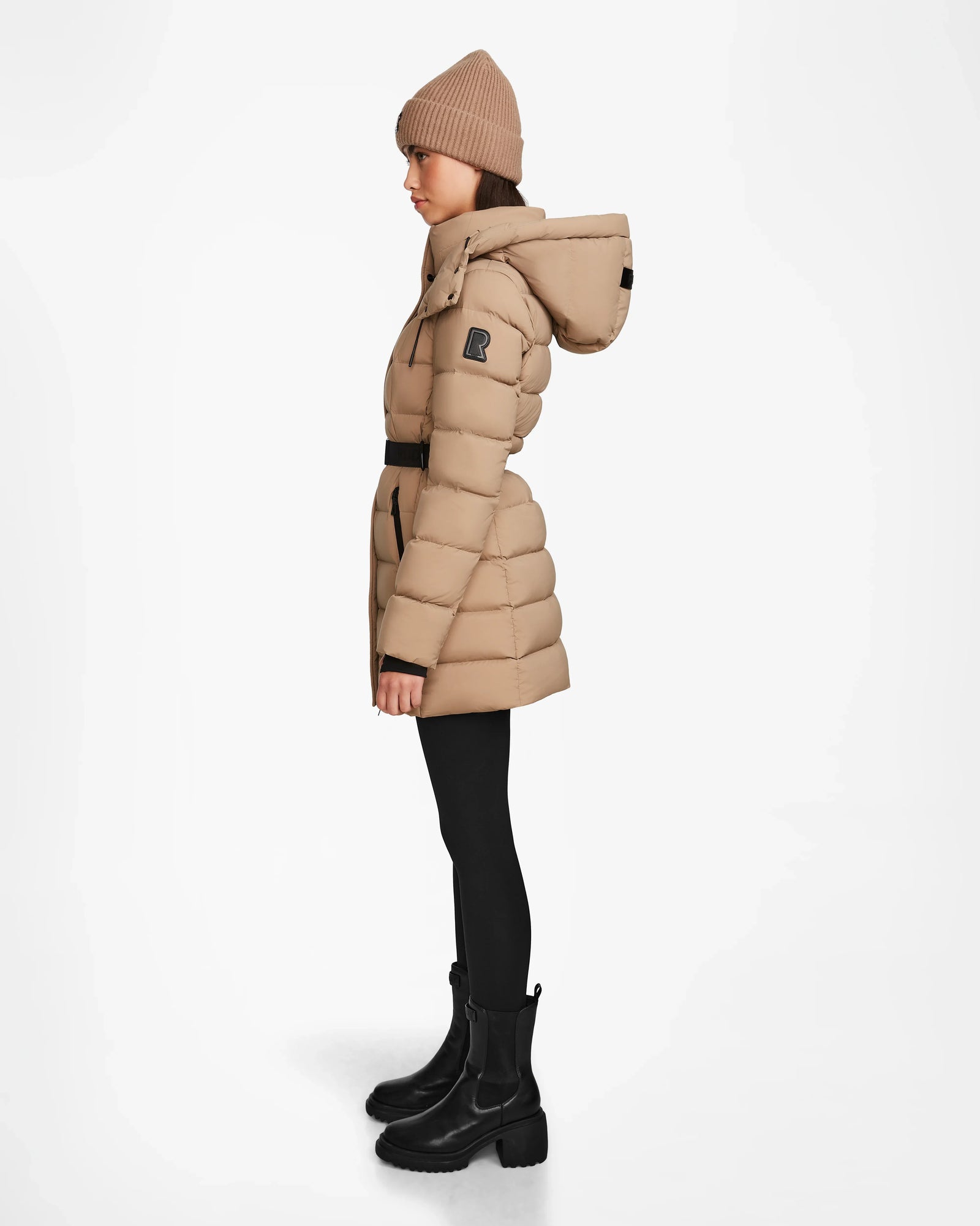 A woman wearing the Rudsak FRIDA M - L124524 Sand quilted down jacket with removable hood, a tan knit beanie, black leggings, and black boots stands in profile against a plain white background.