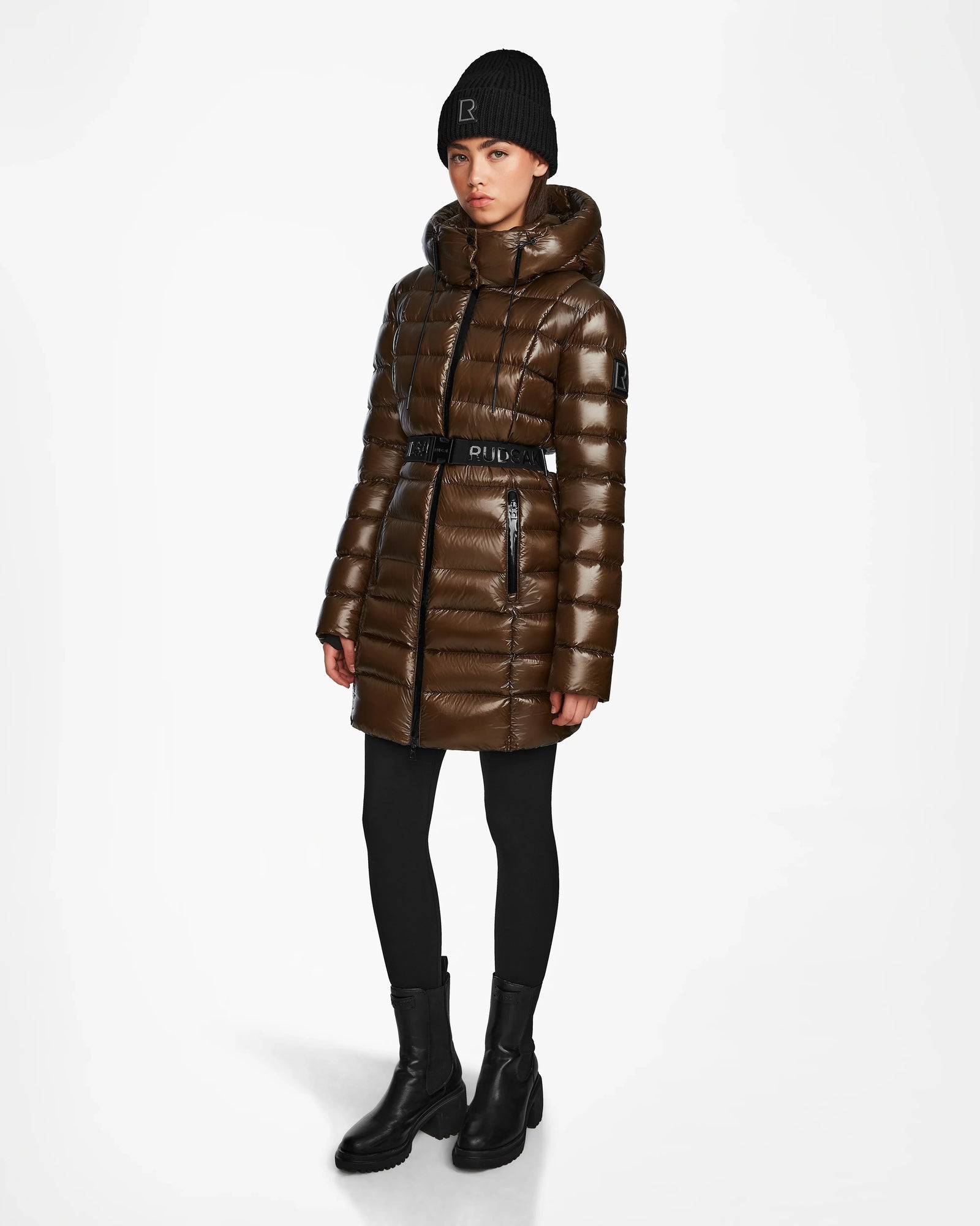 A person wearing the Rudsak FRIDA S - L123513 women's mid-length down jacket in light olive, styled with black leggings, black ankle boots, and a black beanie stands against a plain white background.