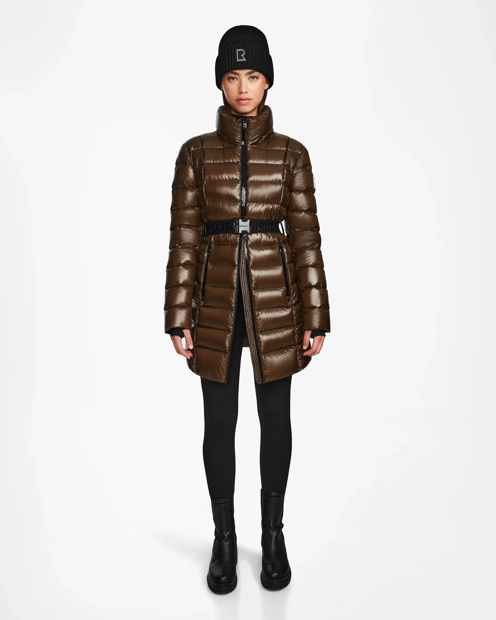 A woman faces forward, wearing the Rudsak FRIDA S - L123513 Light Olive women’s mid-length down jacket with a belted waist and removable hood, paired with black leggings, ankle boots, and a beanie against a plain white background.
