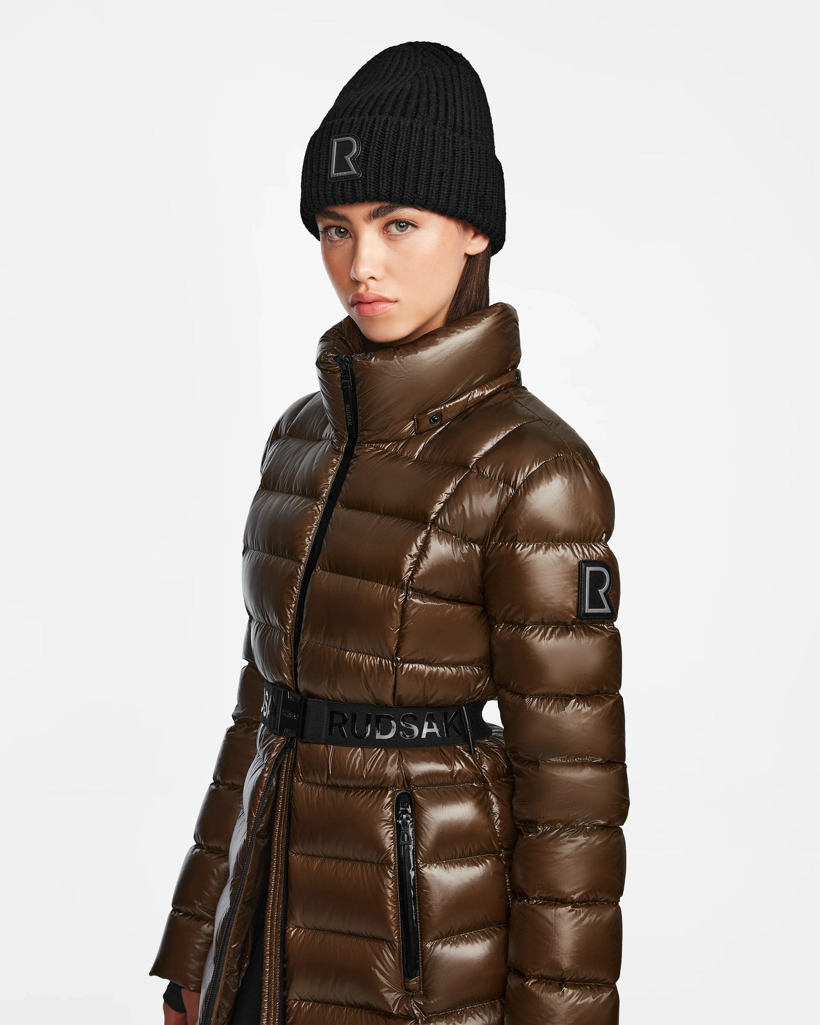A woman wears the Rudsak FRIDA S - L123513 Light Olive women’s mid-length down jacket with a high collar, matching belt, and R logo, paired with a black knit beanie. The quilted jacket stands out against a plain white background.