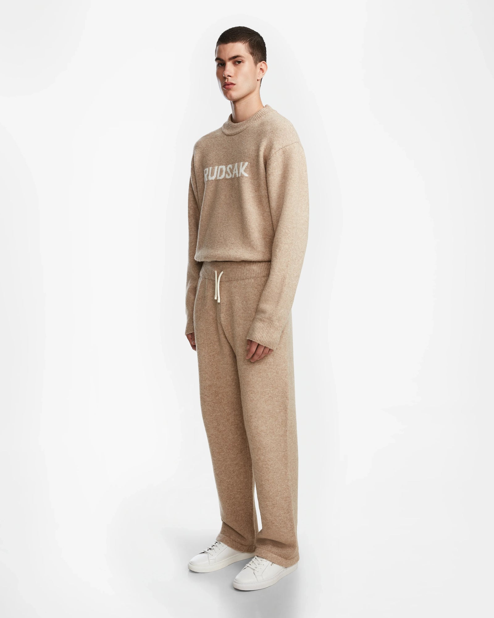 A person wears the Rudsak GEIGER - 9524512 Camel M knit sweater and matching relaxed fit lounge pants with a drawstring, paired with white sneakers, standing against a plain white background.