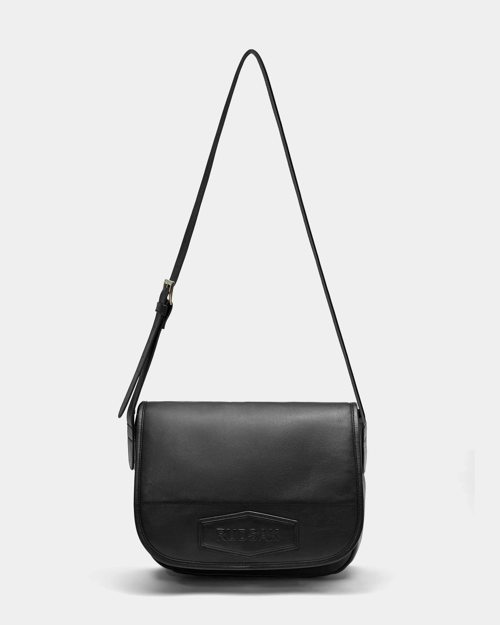 The Rudsak GRAYSON - 8324512 Black is a leather crossbody bag with a long adjustable strap and flap closure, featuring a rectangular embossed logo patch on the front, shown against a plain white background.