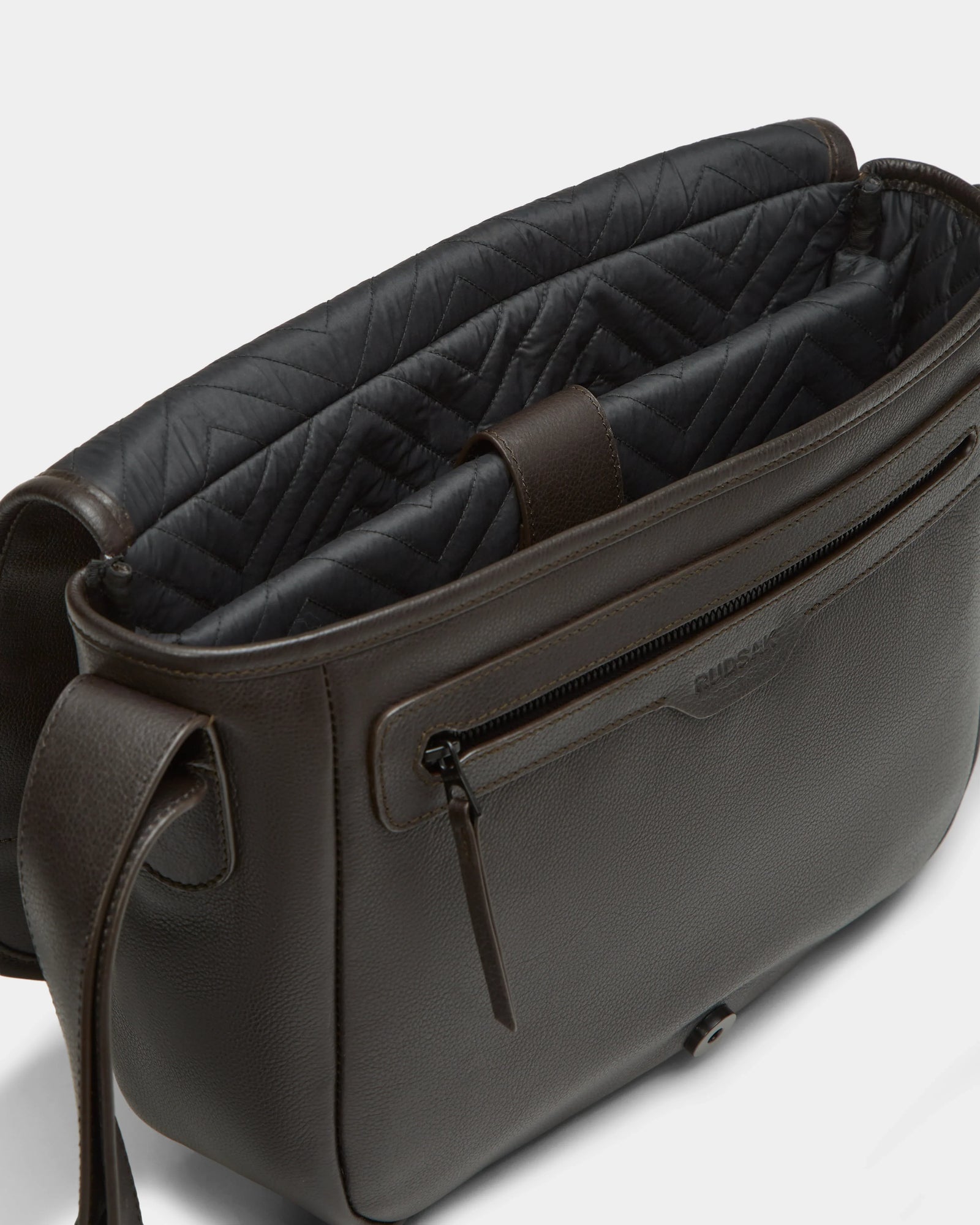 The Rudsak GRAYSON - 8324512 in Espresso is a dark brown leather crossbody bag featuring an adjustable strap, front zip pocket, textured exterior, and black quilted lining, shown open to display its roomy compartments.