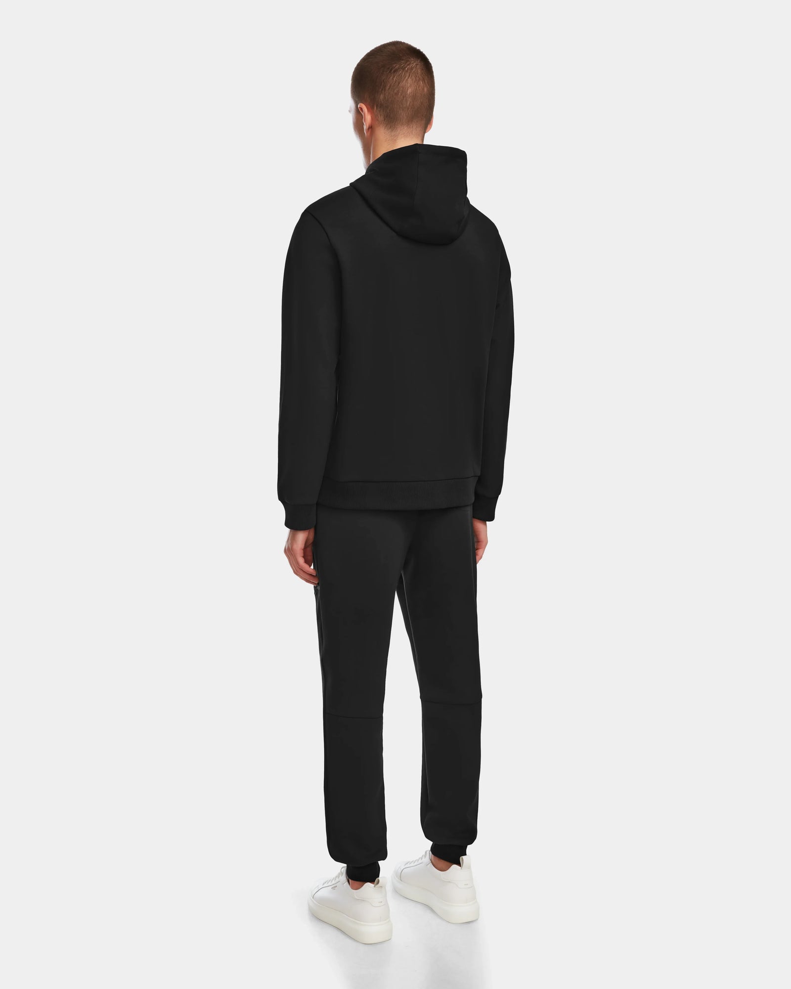 A man stands facing away, wearing the black Rudsak JARED - 6524533 hoodie and matching sweatpants with white sneakers, set against a plain light gray background.