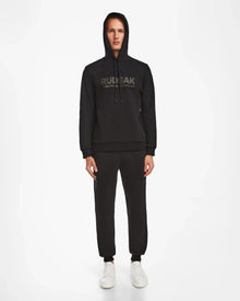 A man faces forward in a black Rudsak JARED - 6524533 hoodie with the hood up, matching jogger pants, and white sneakers. The men’s cotton-blend hoodie features RUDSAK branding on the front. Plain white background.