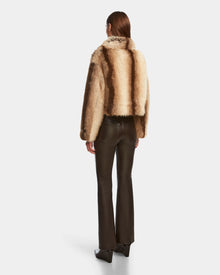 A person with straight brown hair, seen from behind, wears the Rudsak JOSILYN - 8124792 Mocha faux fur jacket—an essential winter look—paired with dark brown leather pants and black shoes against a plain white background.