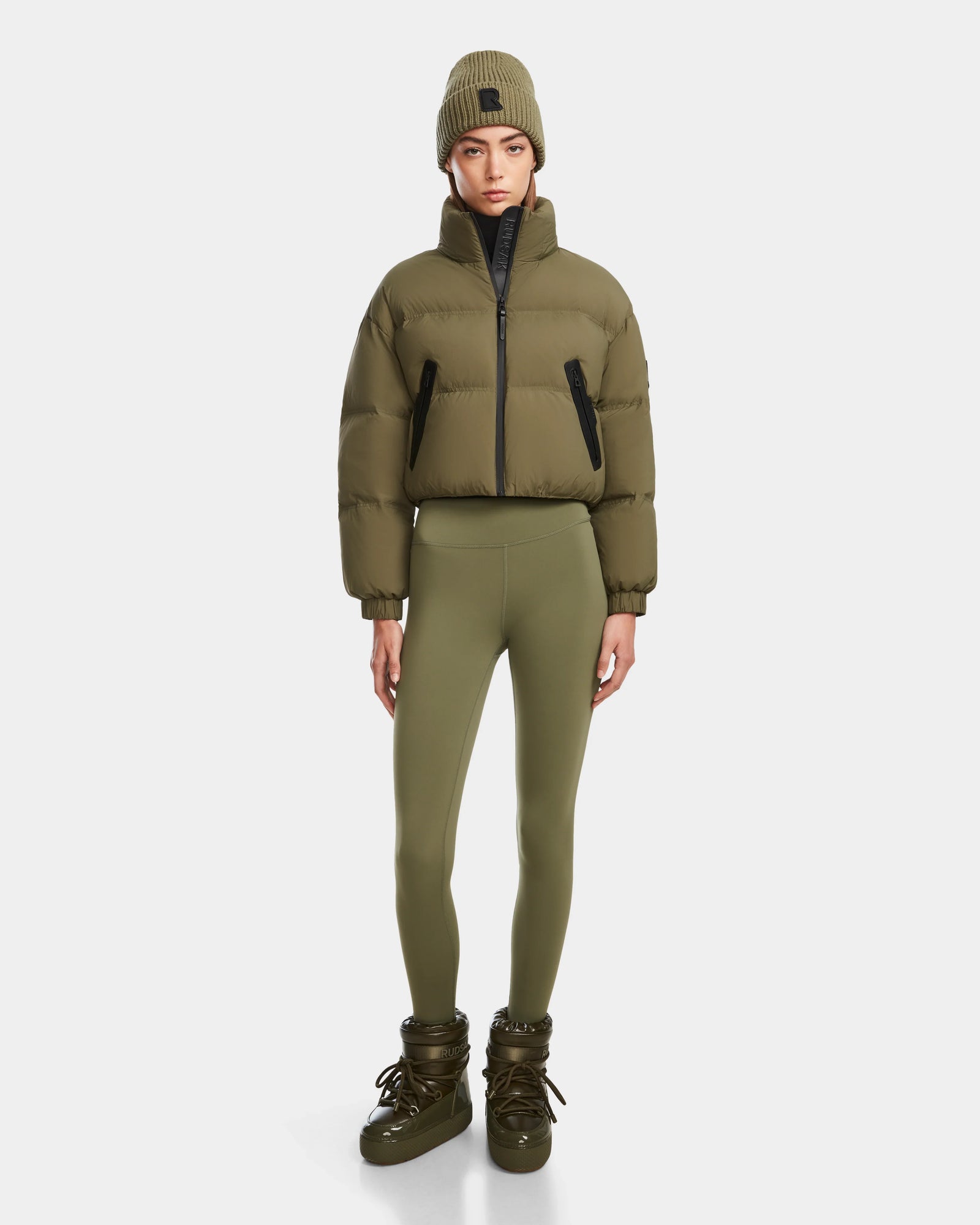 A woman in the Rudsak KARLEE - 8124721 Loden Green cropped down puffer, matching leggings, winter boots, and a knit beanie stands against a plain white background.