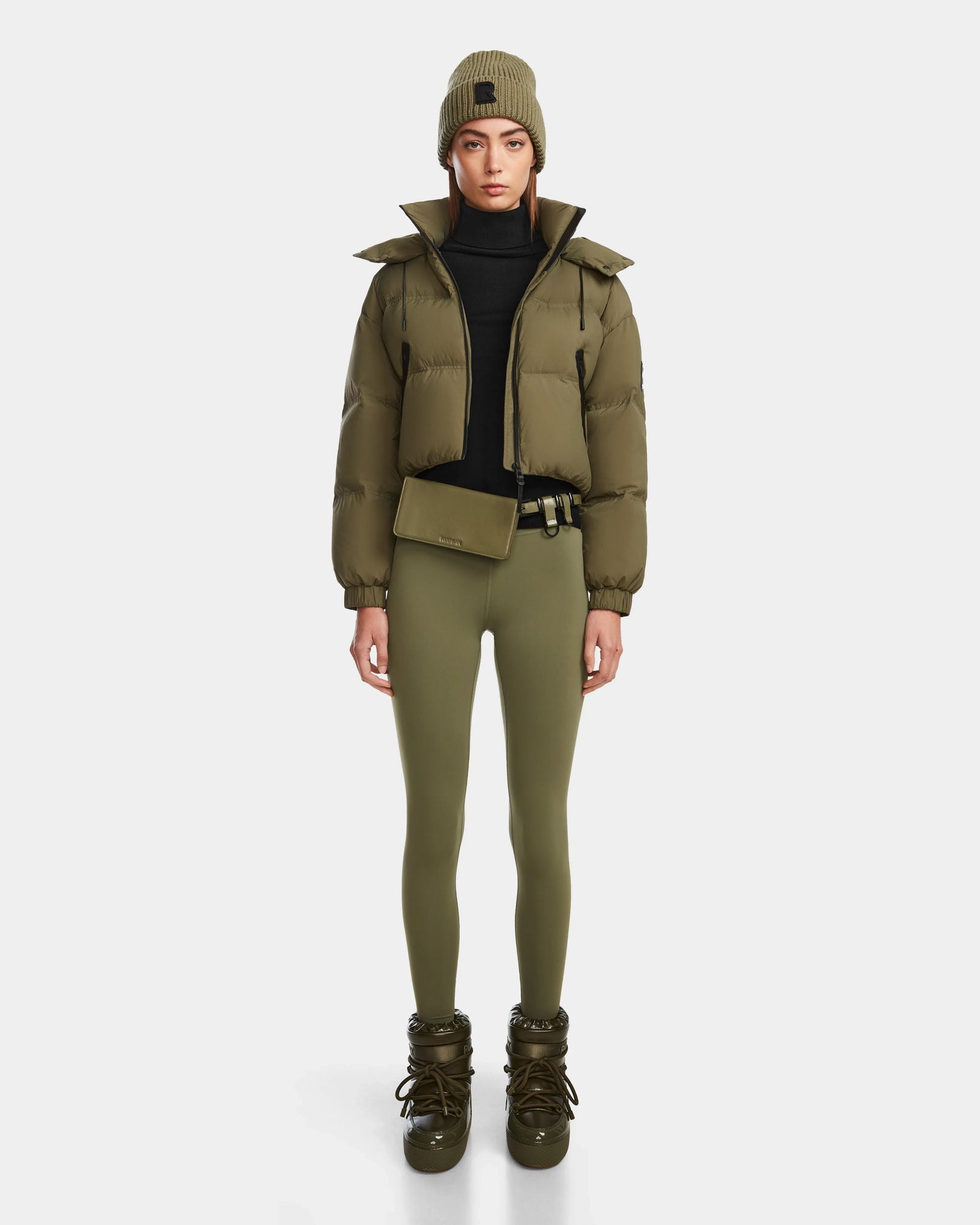 A person faces forward against a plain white background, wearing Rudsak’s KARLEE - 8124721 Loden Green RDS-certified down puffer jacket with matching leggings, a beanie, belt bag, lace-up boots, and a black turtleneck.