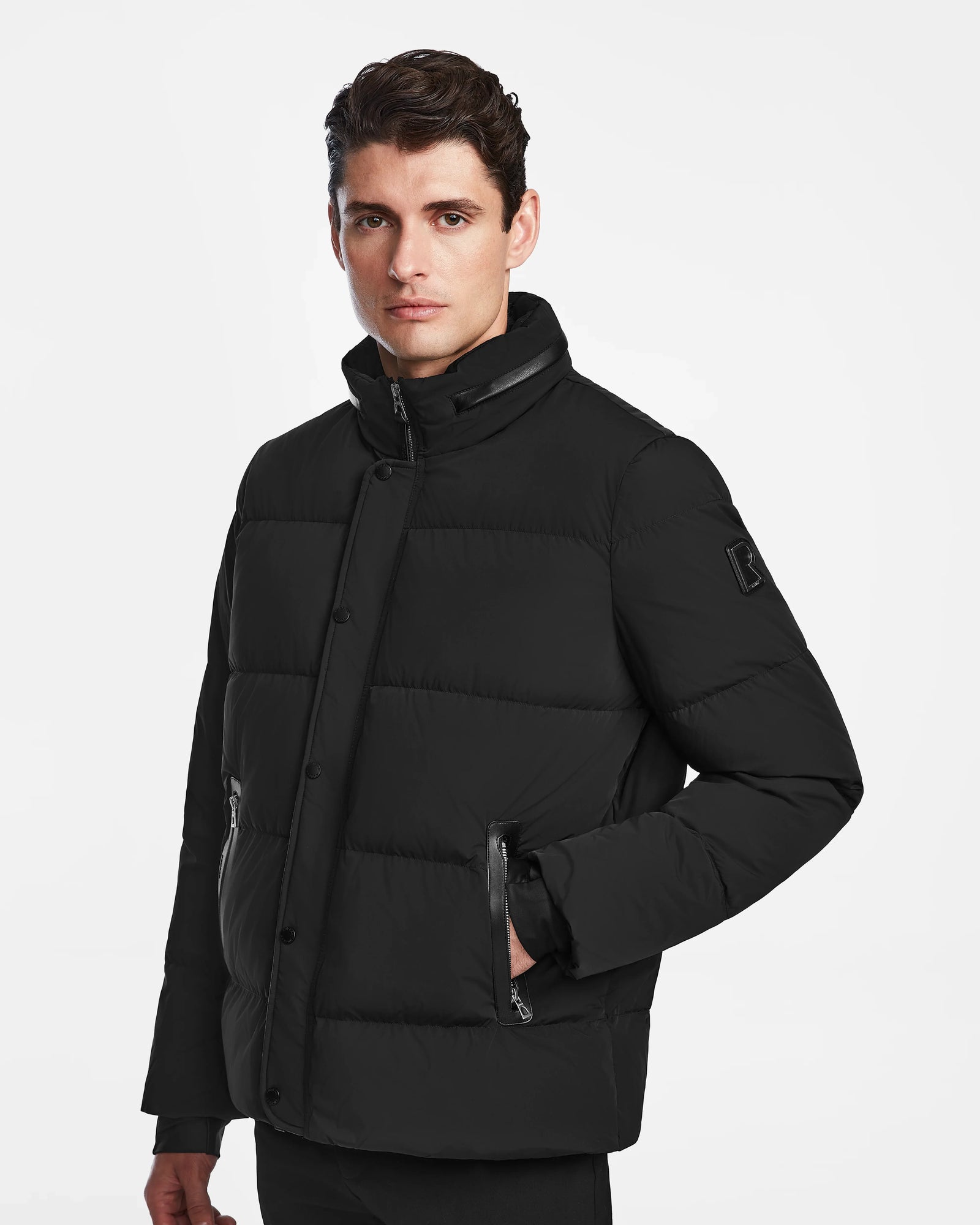 A man with dark hair wears the Rudsak KARLS - M124505 Black mens puffer jacket, featuring a high collar, quilted design, zippered pockets, and snap buttons. He stands against a plain white background, gazing slightly to the side.