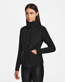 A woman with straight dark hair models the Rudsak KARMA - L124503 Black quilted ecofill jacket featuring a high collar and snap buttons, paired with black pants, posed against a plain white background.