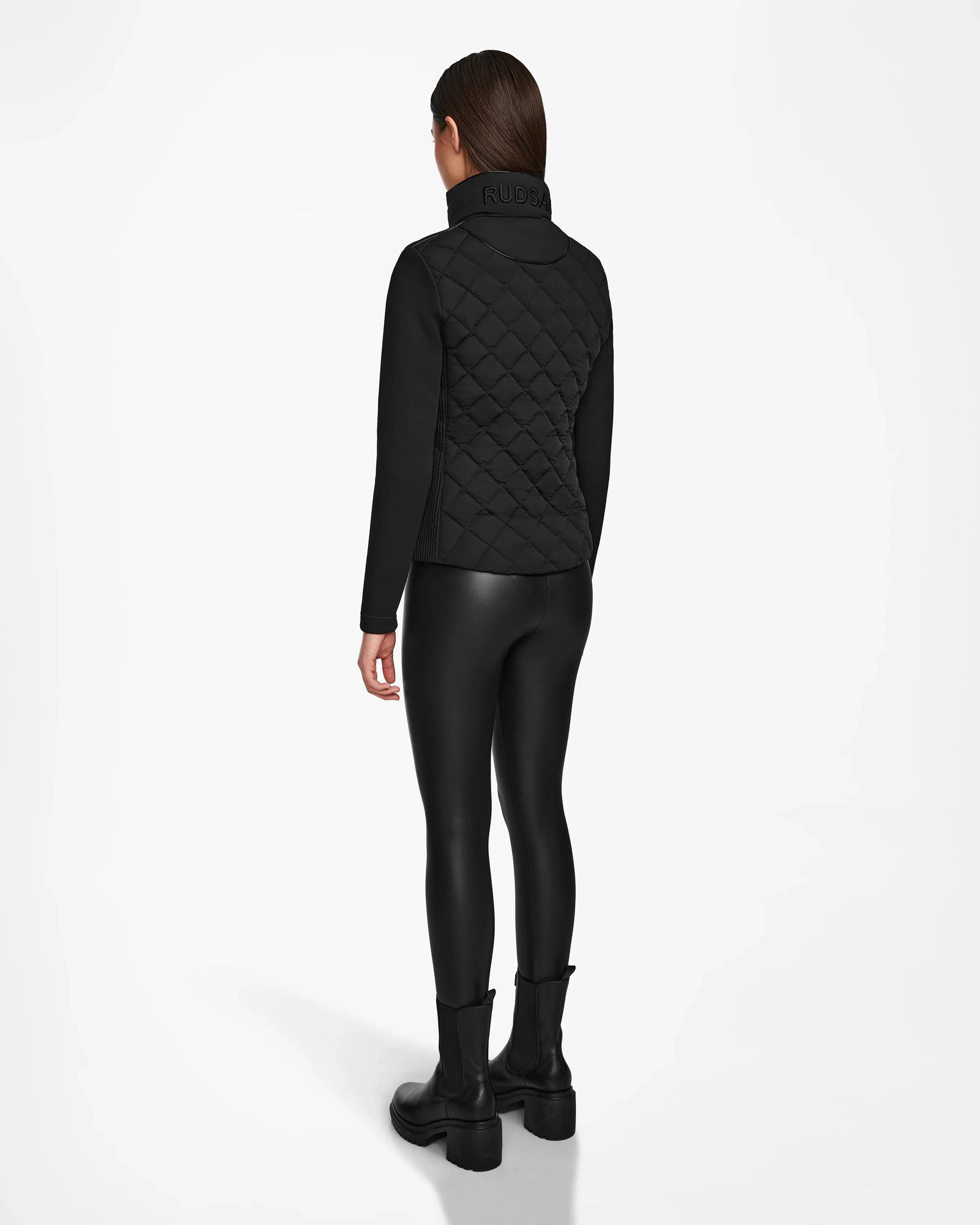 A person faces away, wearing the Rudsak KARMA - L124503 Black fitted womens matte light jacket for sustainable warmth, styled with black leggings and boots. The plain white background highlights this ideal transitional weather look.
