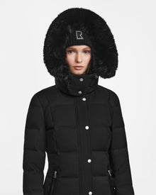 A woman wearing the Rudsak KEHLANI - L124540 black quilted down coat with a large faux fur-trimmed hood and a black beanie stands against a plain white background.