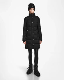 A person faces forward against a plain white background, wearing the Rudsak KEHLANI - L124540 Black long quilted down coat, paired with a black knit beanie, black leggings, and black winter boots.