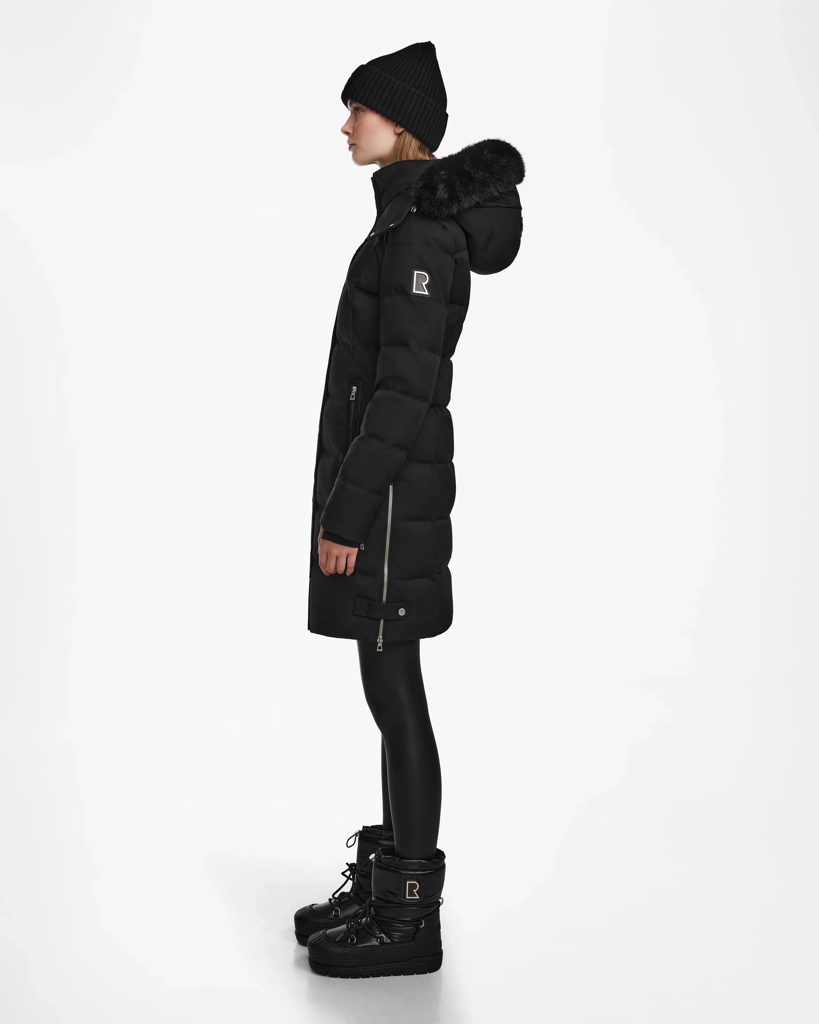 A woman in profile wears the KEHLANI - L124540 Black down coat by Rudsak, featuring a fur-lined hood—styled with black leggings, winter boots, and a knit beanie for an effortlessly chic winter look against a white background.