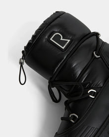 Close-up of the Rudsak KIKA - L224506 Black women’s winter boot featuring padded black material, black laces, silver eyelets, a bold metallic R logo on the upper cuff, and a chunky rubber sole, shown against a plain white background.