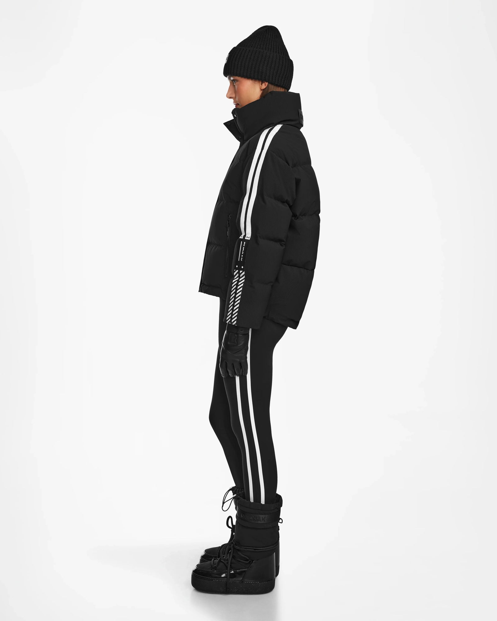 A person in profile wears a black beanie, gloves, winter boots, and the Rudsak KIMIA - 8124716 Black down ski jacket with bold white side stripes over matching leggings, providing stylish warmth against a plain white backdrop.