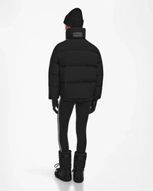Dressed in black winterwear, including the Rudsak KIMIA - 8124716 Black ski jacket, beanie, gloves, leggings, and snow boots, a person stands facing away on a plain white background.