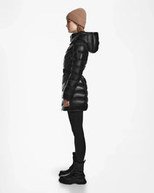 A person in profile wears the Rudsak MALAYA S - L123510 Black women’s down puffer jacket with a hood, paired with black leggings, black boots, and a beige knit beanie, set against a plain white background.