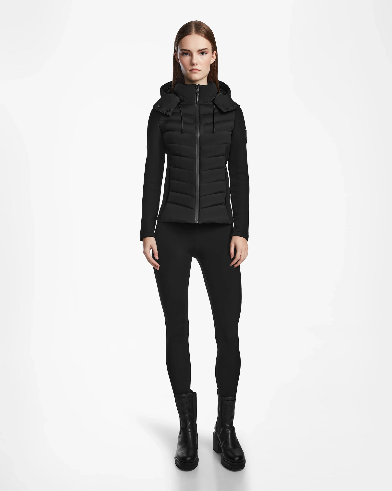 A woman stands facing forward in a Rudsak MALYA M - L123547 Black down quilted jacket, black leggings, and black ankle boots against a plain white background.