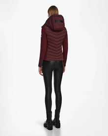 A person stands facing away, wearing the Rudsak MALYA M - L123547 Wine down quilted jacket with a hood, paired with black shiny leggings and black boots against a plain white background.