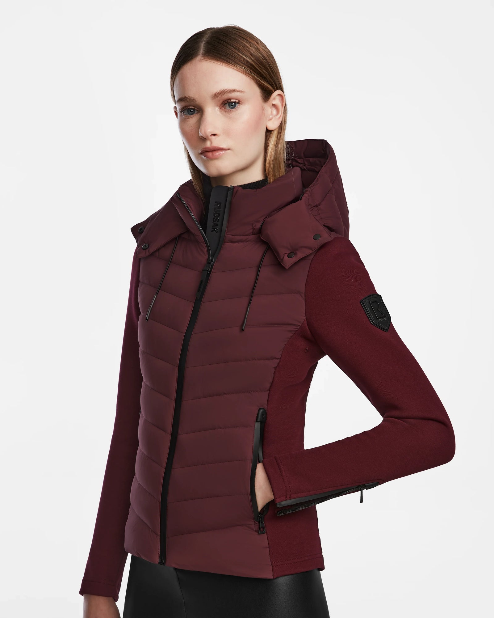 A woman wears the Rudsak MALYA M - L123547 Wine down quilted jacket with a hood, black zippers, and matching long sleeves, standing against a white background.