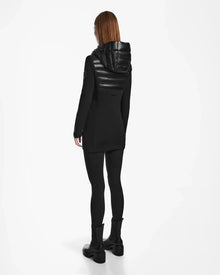 A person with long, straight auburn hair stands facing away, wearing the Rudsak MARRY - L121003 BLACK women’s puffer jacket, black leggings, and black boots against a white background.