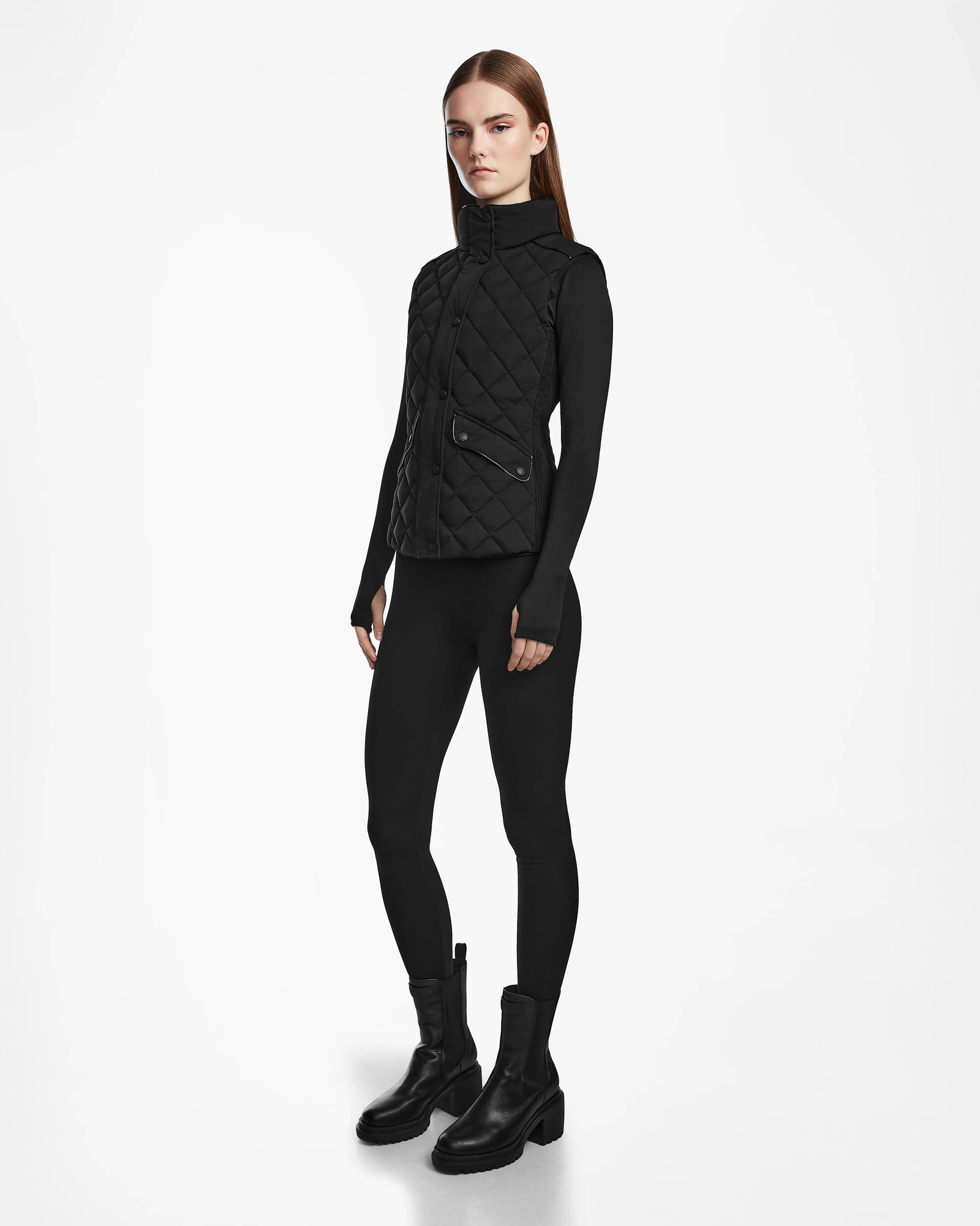 A woman with straight brown hair and a neutral expression models the Rudsak MATILDA - L124504 Black vest over a black long-sleeve top, leggings, and boots against a white background—an effortlessly chic transitional look.