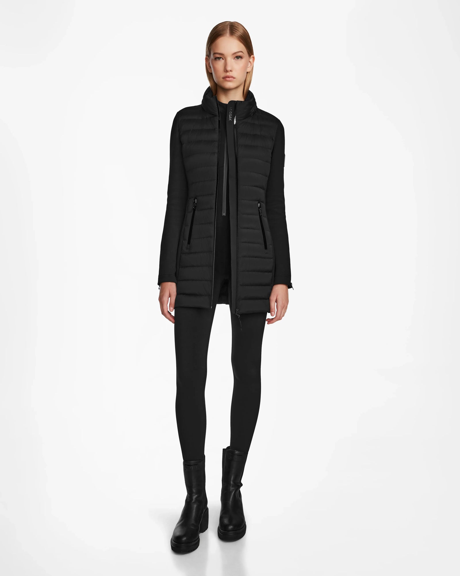 A woman stands facing forward wearing the RUDSAK MERINA - 8122546 BLACK long quilted down jacket over black leggings and ankle boots against a plain white background.