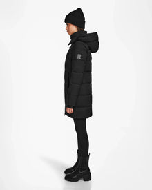 Wearing the Rudsak MIRIAM - L122523 BLACK weather-resistant puffer coat with black beanie, pants, and boots, this look in profile against a white background is ideal for any winter wardrobe.