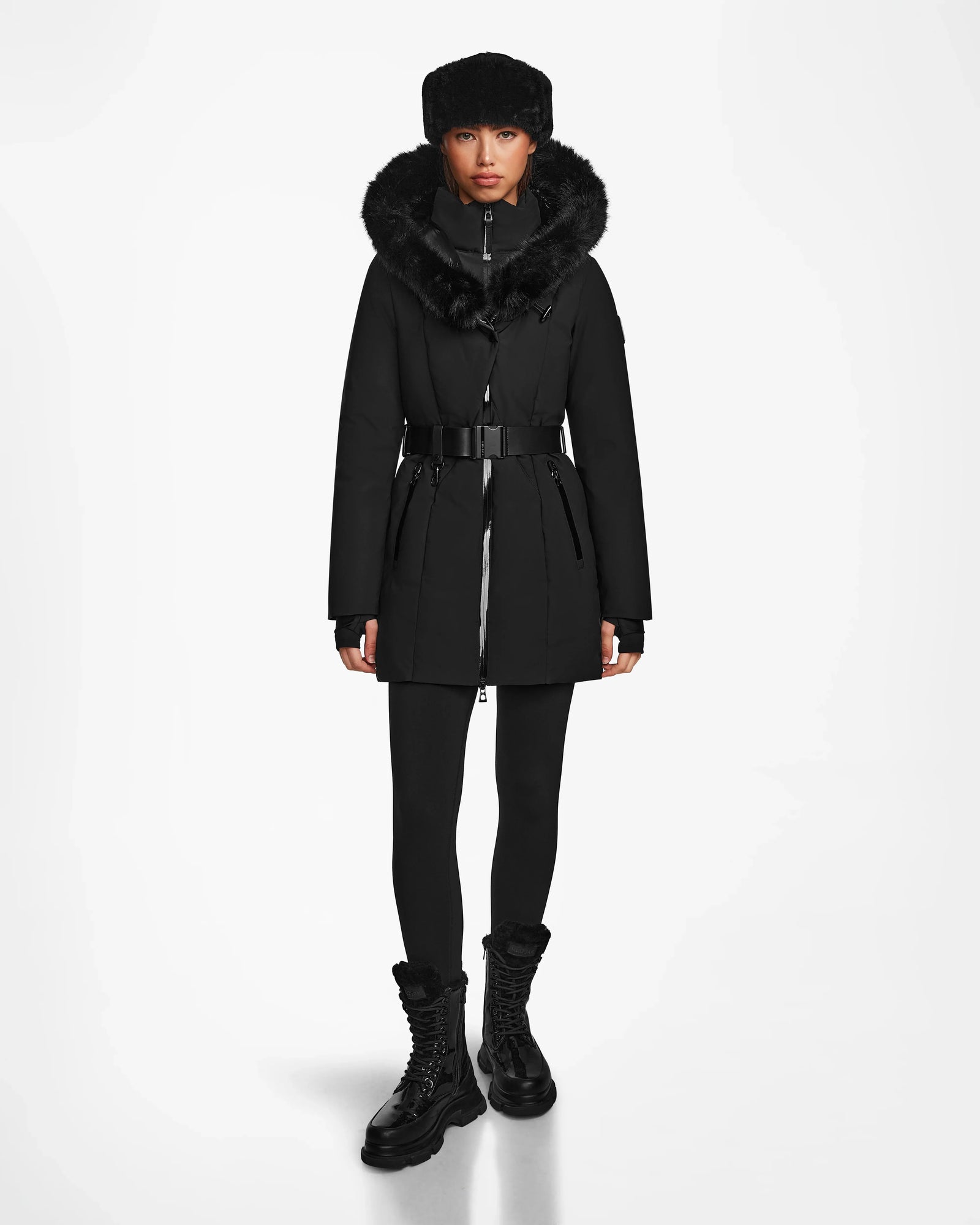 A woman faces forward in the Rudsak MODA M - 8124555 Black down parka with a fur-lined hood, black leggings, lace-up boots, and a matching fur hat. The fitted coat creates a stylish silhouette against a plain white background.