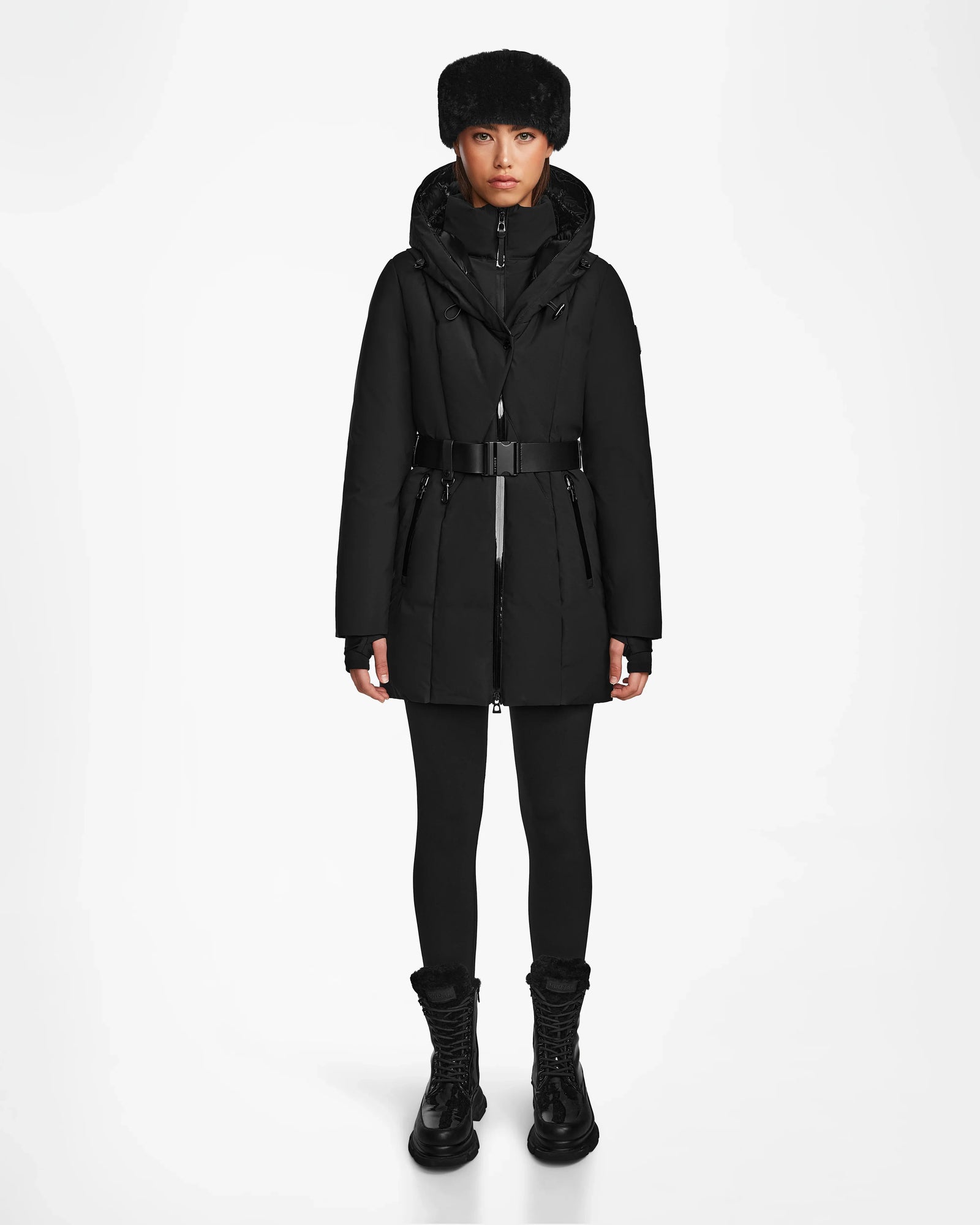 A person stands facing forward in the Rudsak MODA M - 8124555 Black winter coat with belt, black leggings, boots, and a fur hat against a white background—showcasing timeless style and sustainable design.