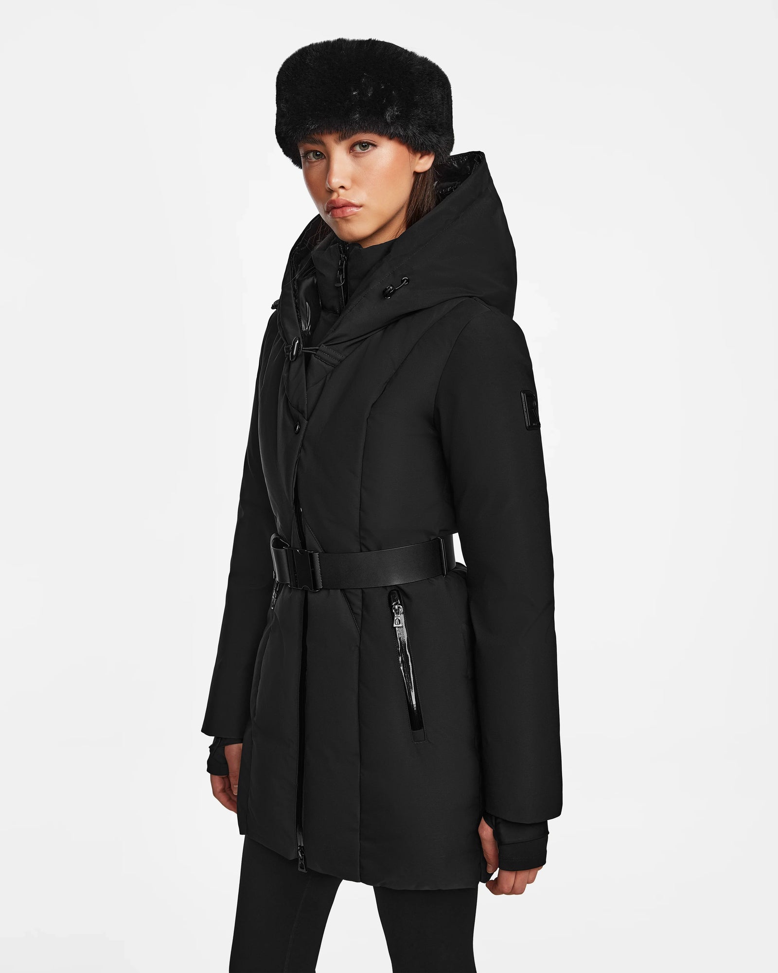 A woman showcases sleek winter style wearing the Rudsak MODA M - 8124555 Black belted coat with a high collar and a black faux fur hat against a plain white background.