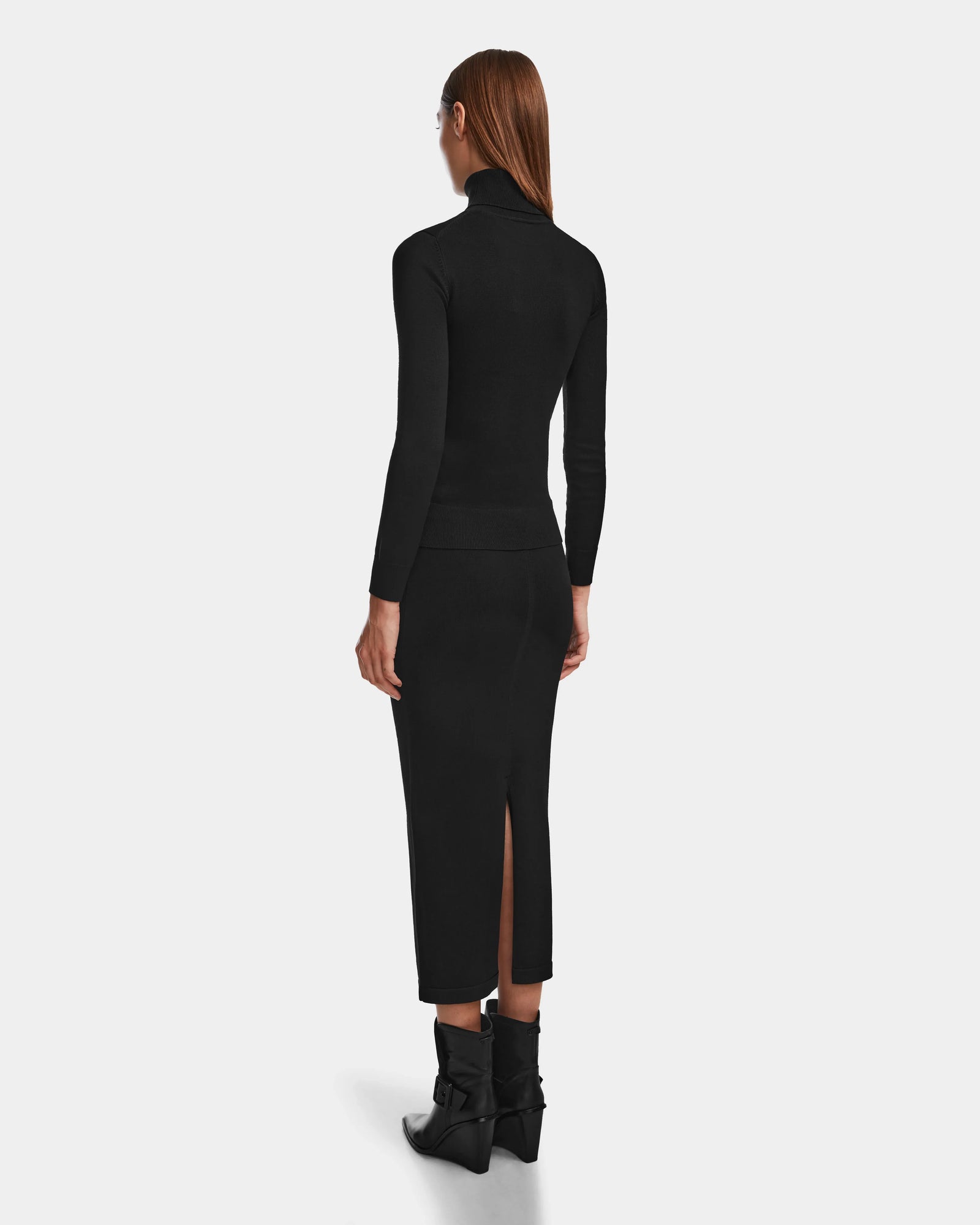 A woman with straight, long brown hair is seen from behind in a fitted black turtleneck and the MONICA - 8524516 Black knit skirt by Rudsak, which features a back slit. She pairs it with black ankle boots with bows against a plain white background.