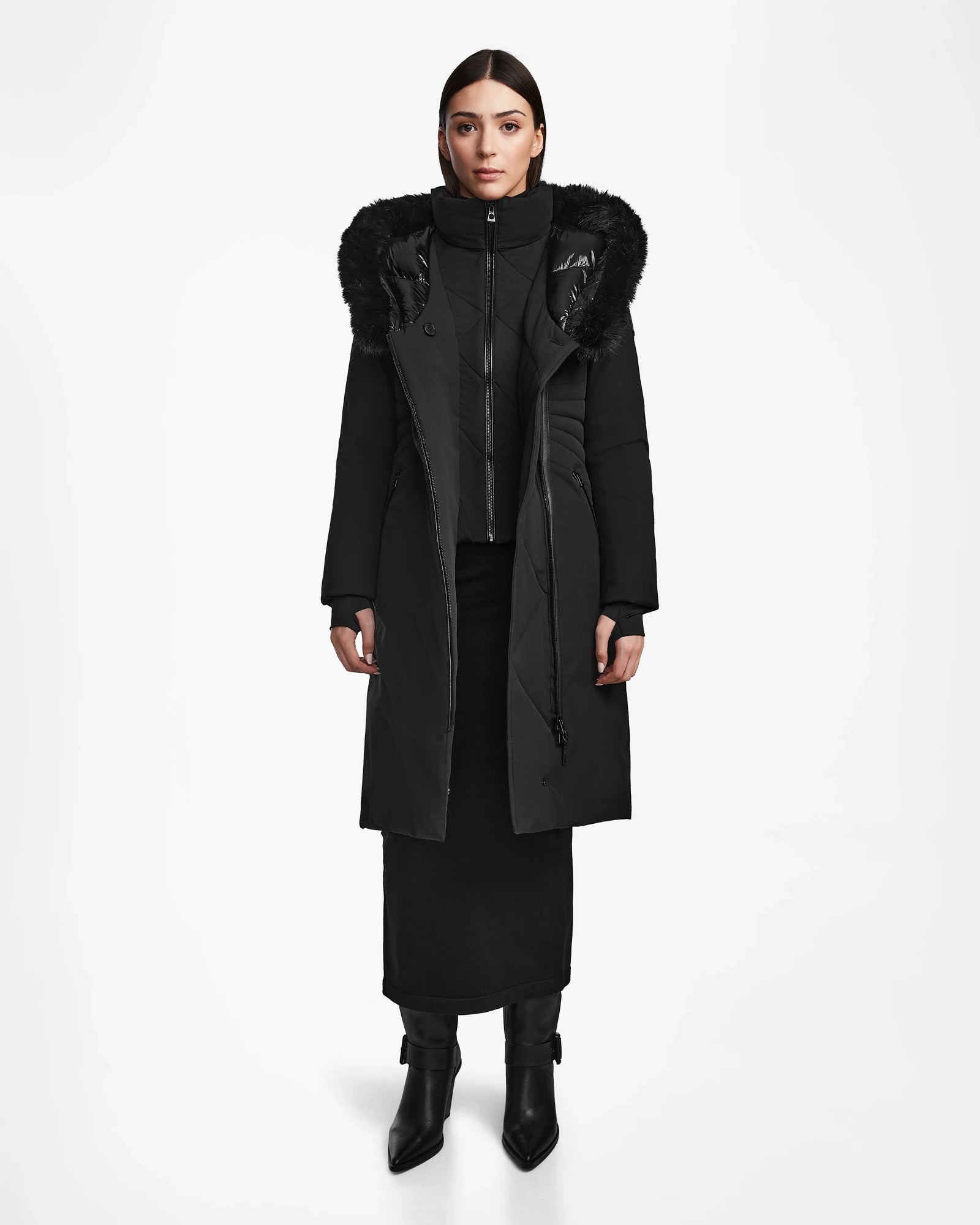 A woman faces forward wearing the Rudsak MUSA - L124534 Black long down puffer with a fur-trimmed hood, paired with a black skirt, black boots, and a high-neck layer, set against a plain white background.