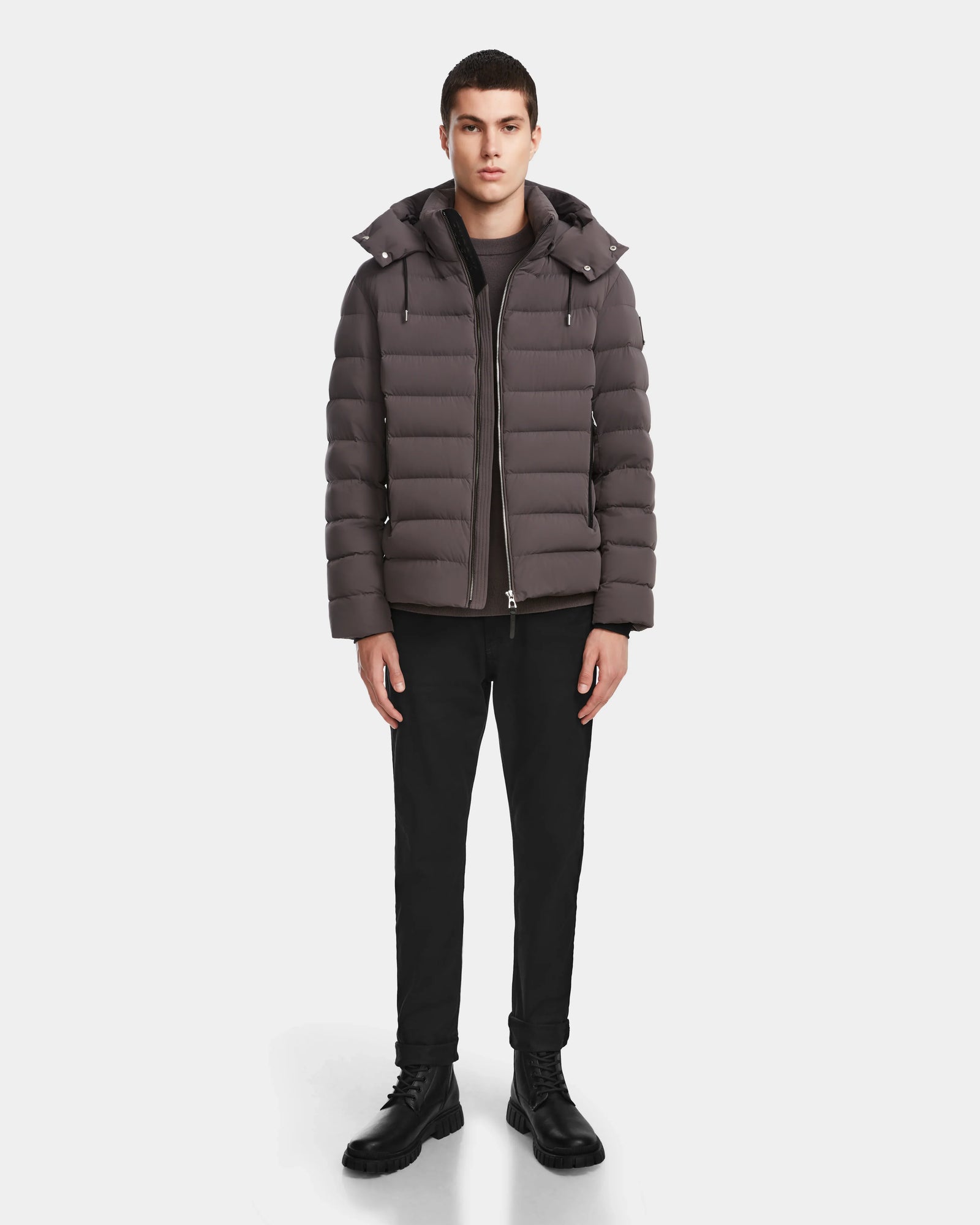 A man faces forward wearing the Rudsak NAVAN - 6124551 Grey mens quilted down puffer, wind and water-repellent, paired with black pants and black boots against a plain white background.