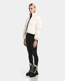A woman wears the Rudsak OPRAH - 8124708 Iceland cropped down puffer, styled with black leggings and black winter boots with white fur trim, standing against a plain light gray background.