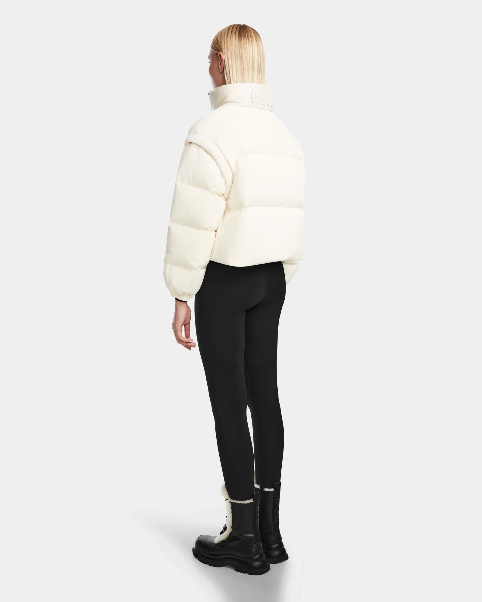 A person with straight blonde hair wears the Rudsak OPRAH - 8124708 Iceland women’s cropped down puffer jacket with removable sleeves, teamed with black leggings and boots, standing facing away from the camera against a plain light background.