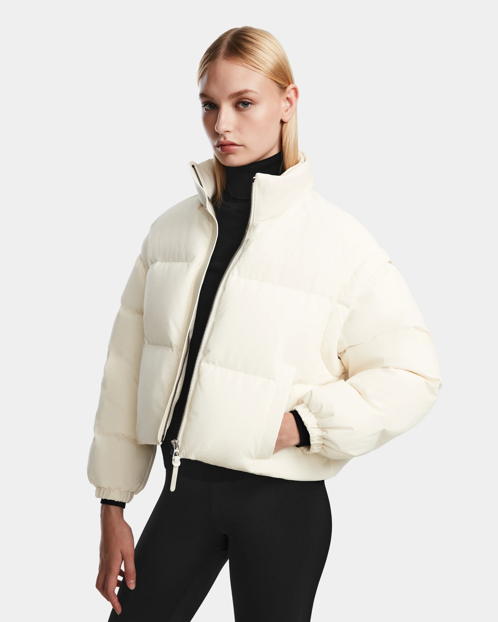 A woman with straight blonde hair wears the cream-colored Rudsak OPRAH - 8124708 Iceland women’s cropped down puffer jacket over a black turtleneck and pants, standing against a plain light gray background.