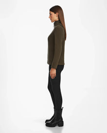 A woman stands in profile facing left, wearing the Rudsak ORCHID - 8524616 Army fitted half-zip pullover sweater with black leggings and chunky boots, against a plain white background.